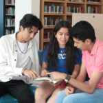 Three teenagers studying together in a library, engaged in a book discussion.