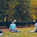 Three women meditate outdoors in autumn park for relaxation and wellness.