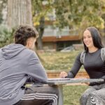 Two young adults sitting at an outdoor table, smiling and engaging in conversation in a park setting.
